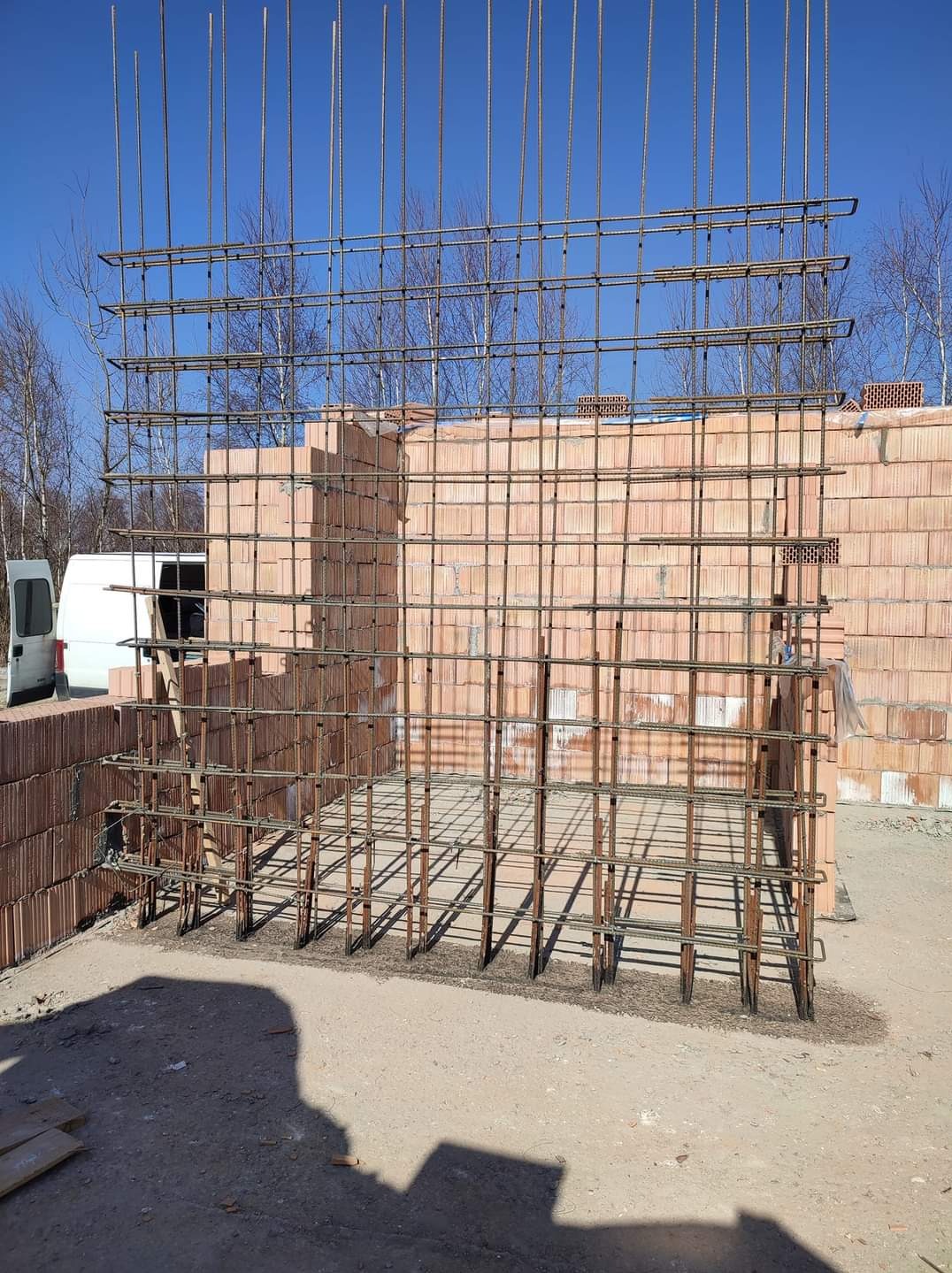 Zbrojenie pionowe i poziome ścian z bloczków ceramicznych, przygotowane do zalania betonem, widok na plac budowy z samochodem dostawczym w tle.