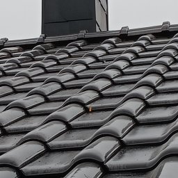 Marcin Dom - Błyszczący, mokry dach z czarnej dachówki ceramicznej z widocznym kominem wentylacyjnym na tle pochmurnego nieba. Na dachu widoczny mały fragment liścia.