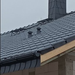 Marcin Dom - Ciemna dachówka ceramiczna na dachu domu w trakcie budowy, widoczny komin z daszkiem, orynnowanie i fragment ściany z pustaków.