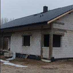 Marcin Dom - Surowy stan budynku z białych bloczków, z czarnym dachem, widoczne otwory okienne i komin, teren wokół niezagospodarowany.