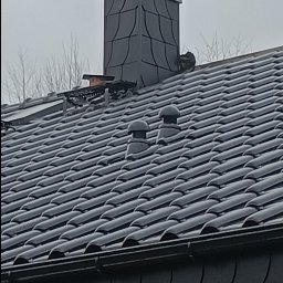 Marcin Dom - Nowy dach pokryty ciemną dachówką ceramiczną, widoczny komin z ozdobnym wykończeniem oraz dwa odpowietrzniki dachowe, całość na tle pochmurnego nieba.