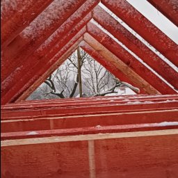 Marcin Dom - Czerwona więźba dachowa w trakcie montażu, pokryta lekkim śniegiem, widok na zaśnieżone drzewa w tle.