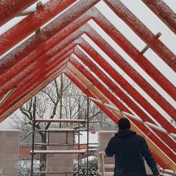 Marcin Dom - Czerwona więźba dachowa pokryta śniegiem, widok od dołu, z pracownikiem w tle i ośnieżonymi bloczkami oraz rusztowaniem w oddali.