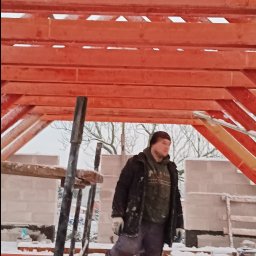 Marcin Dom - Konstrukcja drewniana dachu w trakcie budowy, pokryta śniegiem, z widocznym pracownikiem w kurtce i czapce, stojącym na belce, w tle ściana z bloczków.