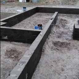 Marcin Dom - Wylane fundamenty domu z hydroizolacją pionową, widoczny teren budowy z narzędziami i pryzmą piasku w tle.