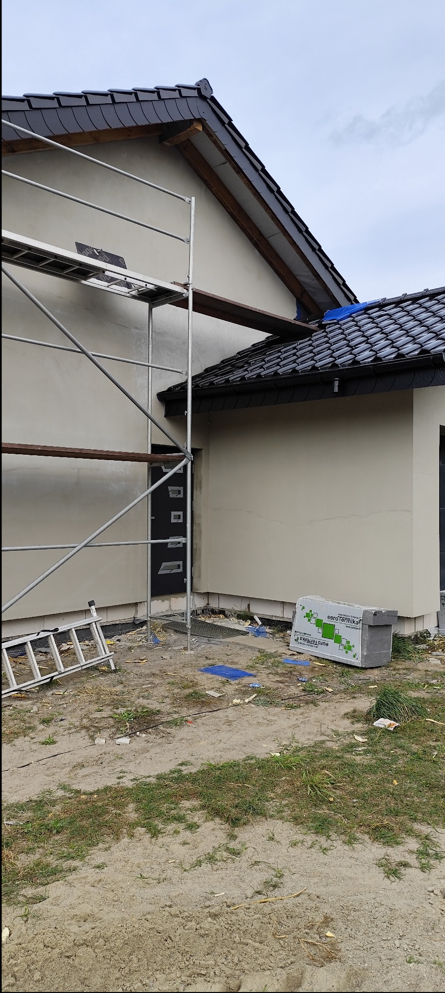 Narożnik domu w trakcie ocieplania elewacji, widoczne rusztowanie, drabina, paczka materiałów izolacyjnych i fragment dachu z czarną dachówką.