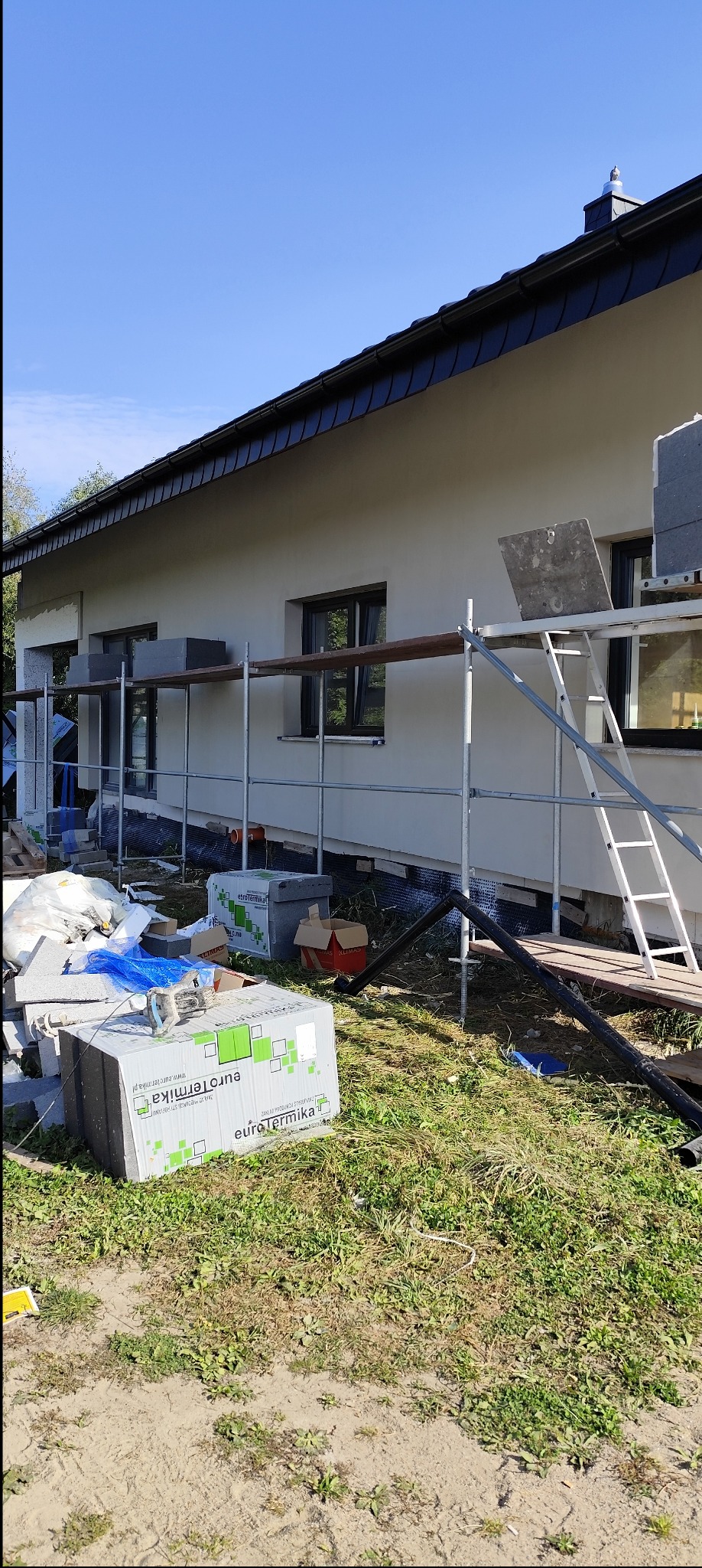 Budynek jednorodzinny w trakcie ocieplania, widoczne rusztowania, materiały izolacyjne w opakowaniach Eurotermika oraz częściowo ocieplona ściana boczna z oknami.