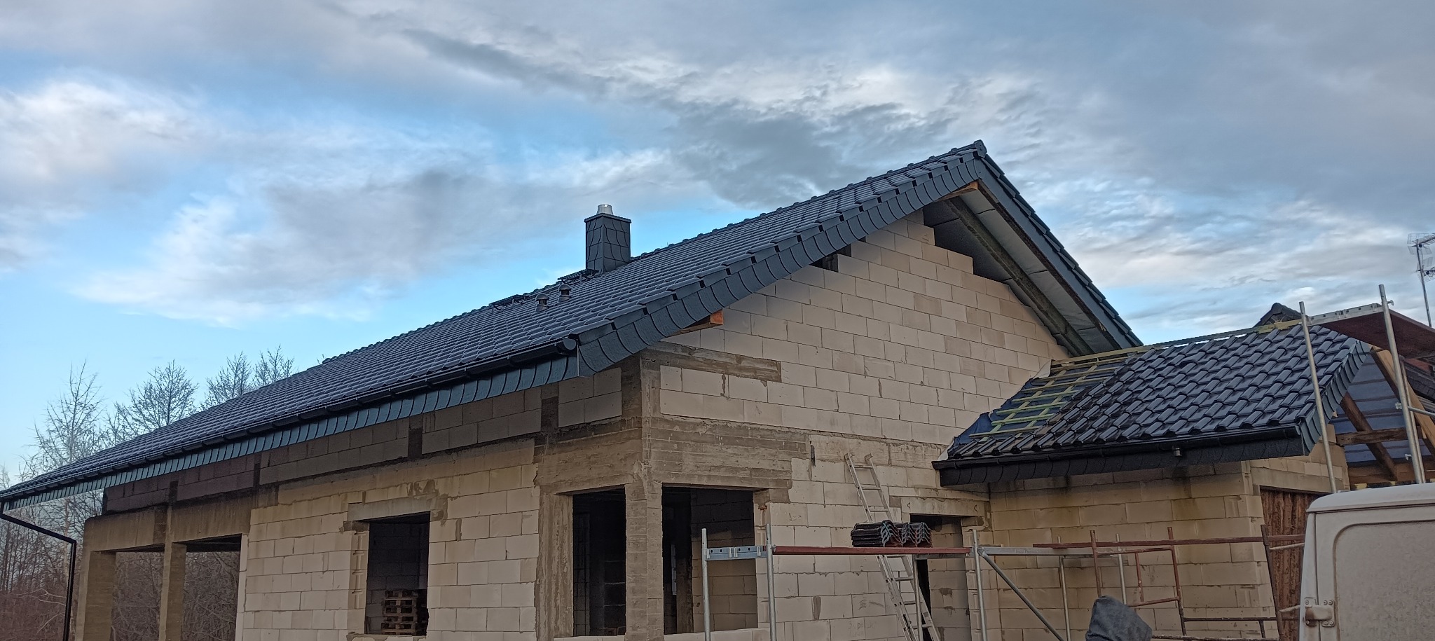 Budowa domu murowanego z widoczną czarną dachówką na ukończonym fragmencie dachu i kominem, ściany z pustaków, rusztowania i drabina w tle, pochmurne niebo.