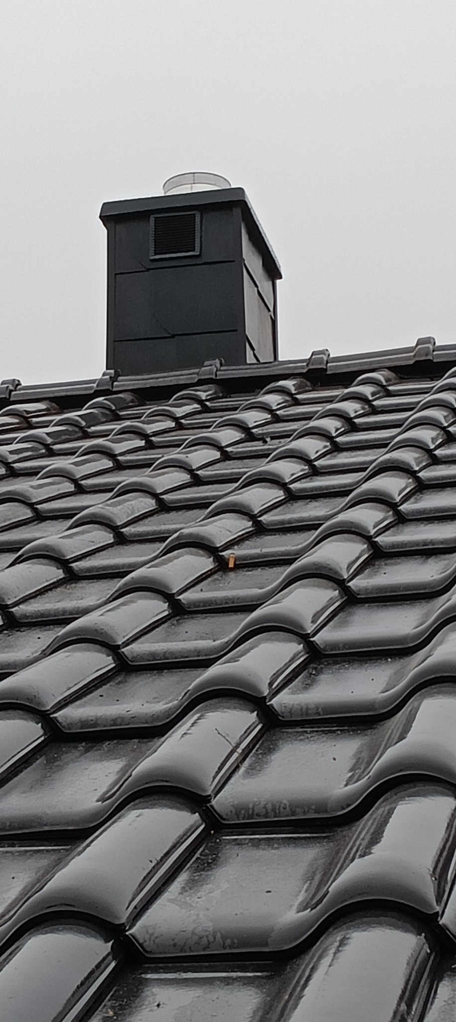 Błyszczący, mokry dach z czarnej dachówki ceramicznej z widocznym kominem wentylacyjnym na tle pochmurnego nieba. Na dachu widoczny mały fragment liścia.