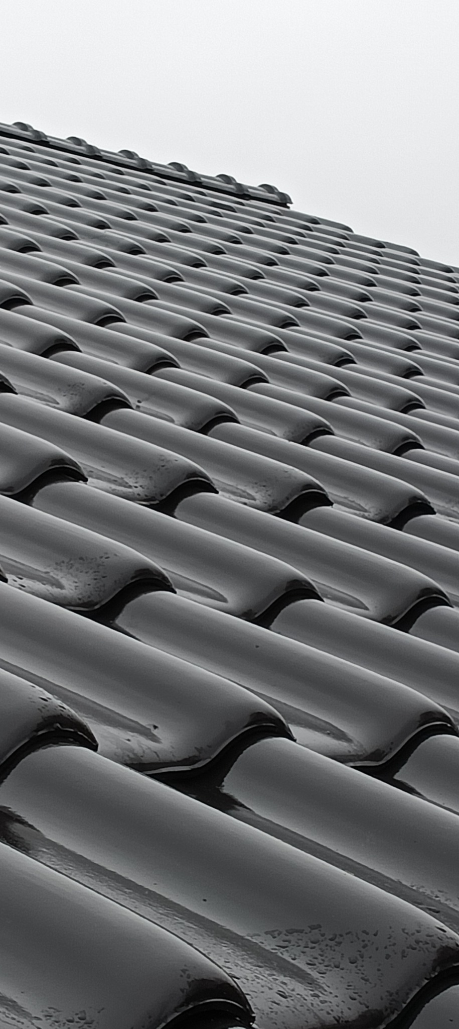 Błyszczące, czarne dachówki ceramiczne ułożone rzędami na dachu, widoczne krople deszczu.