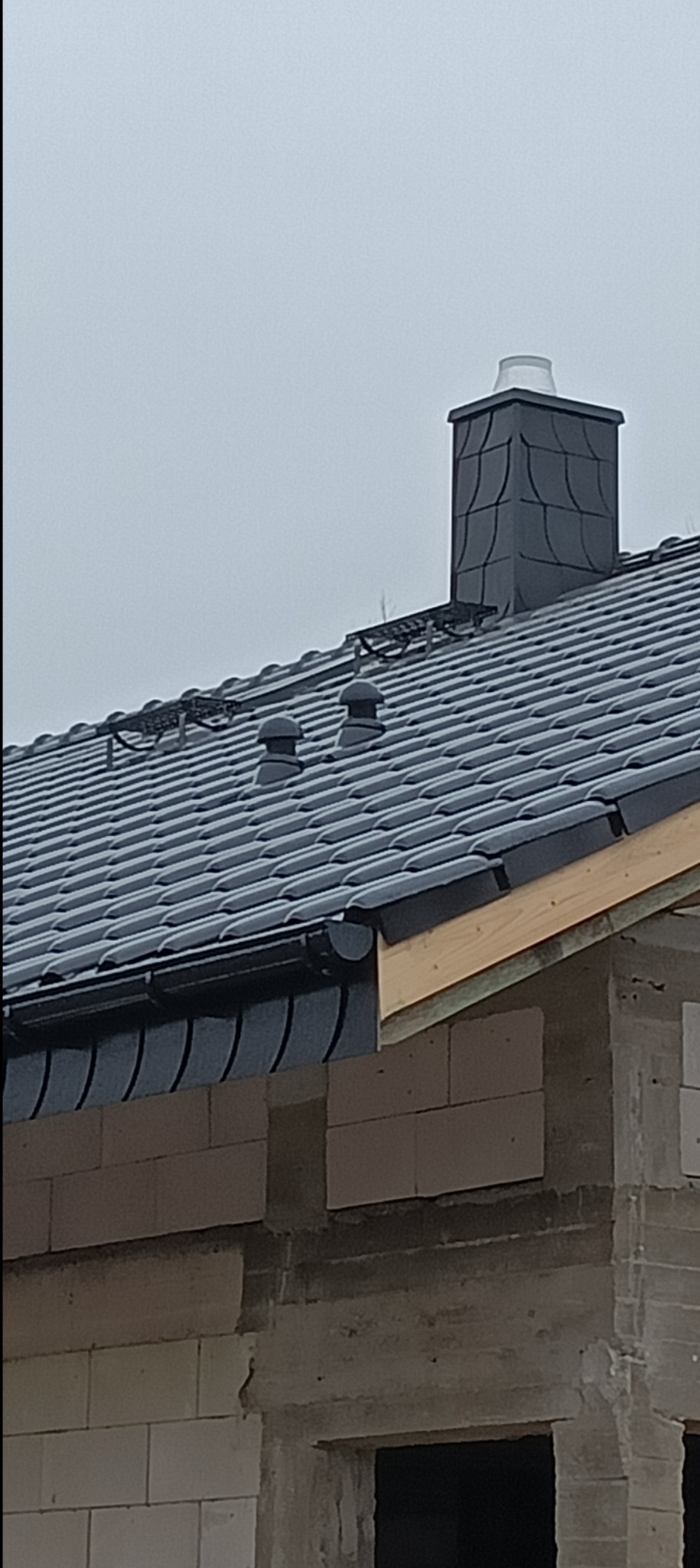 Ciemna dachówka ceramiczna na dachu domu w trakcie budowy, widoczny komin z daszkiem, orynnowanie i fragment ściany z pustaków.