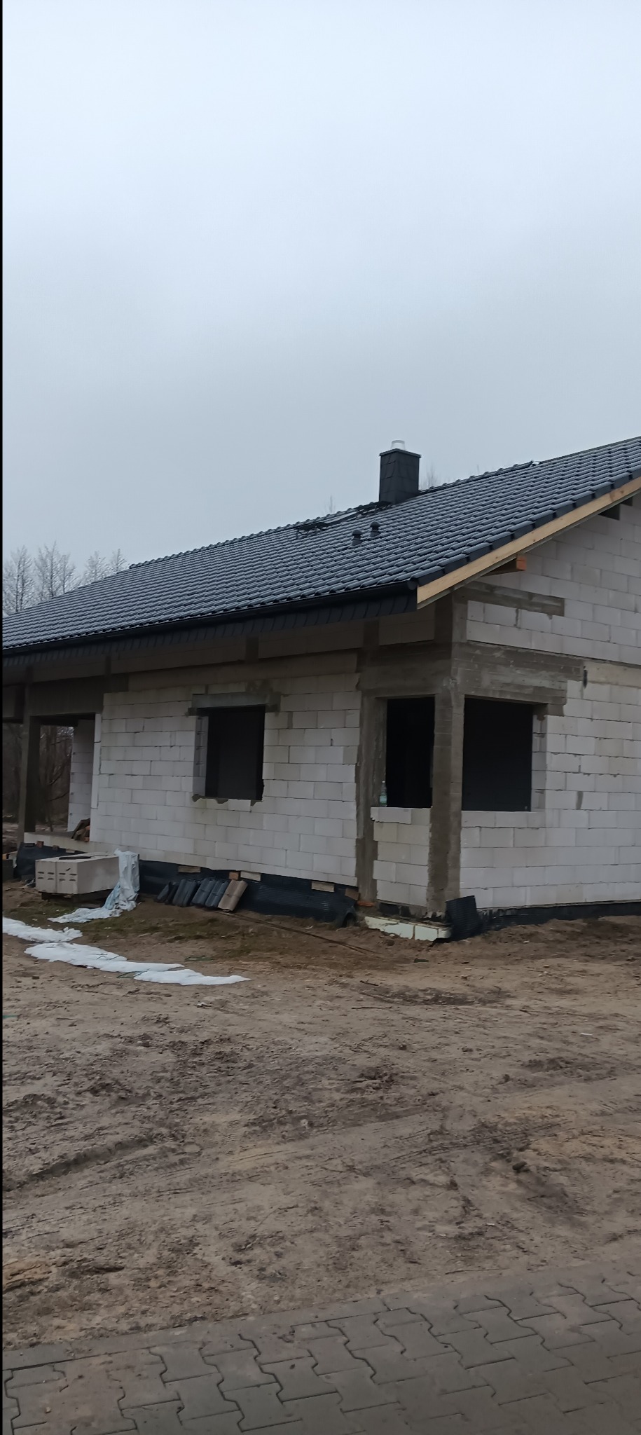 Surowy stan budynku z białych bloczków, z czarnym dachem, widoczne otwory okienne i komin, teren wokół niezagospodarowany.