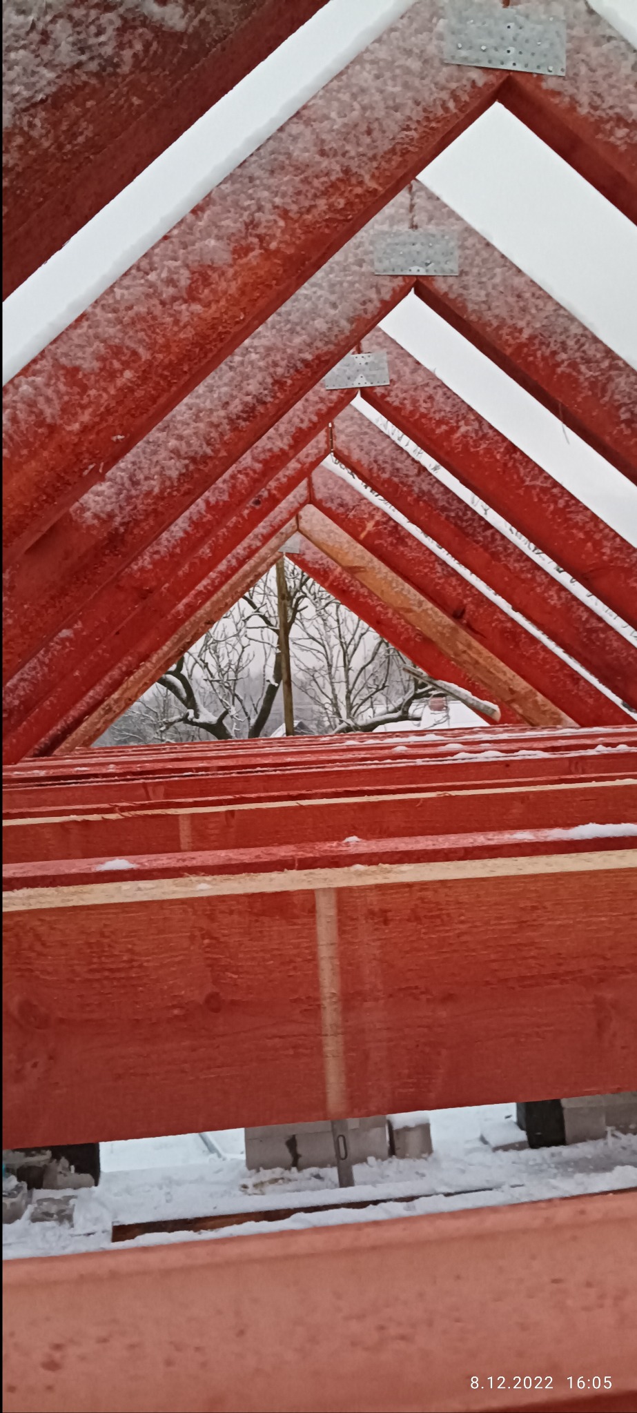 Czerwona więźba dachowa w trakcie montażu, pokryta lekkim śniegiem, widok na zaśnieżone drzewa w tle.