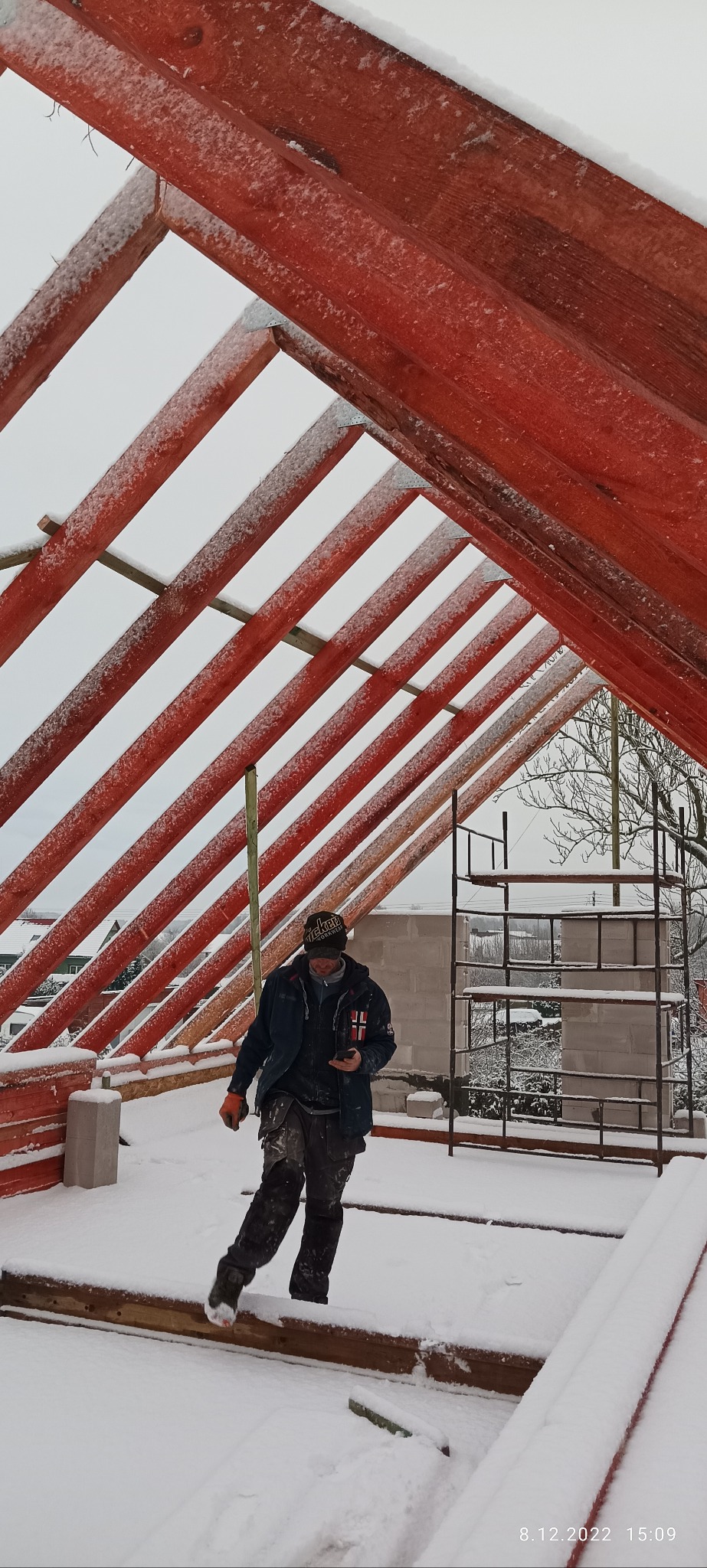 Pracownik budowlany w Świerznie sprawdza postęp prac nad czerwoną, drewnianą konstrukcją dachu pokrytą śniegiem, na tle częściowo wymurowanej ściany i rusztowania w zimowej scenerii.