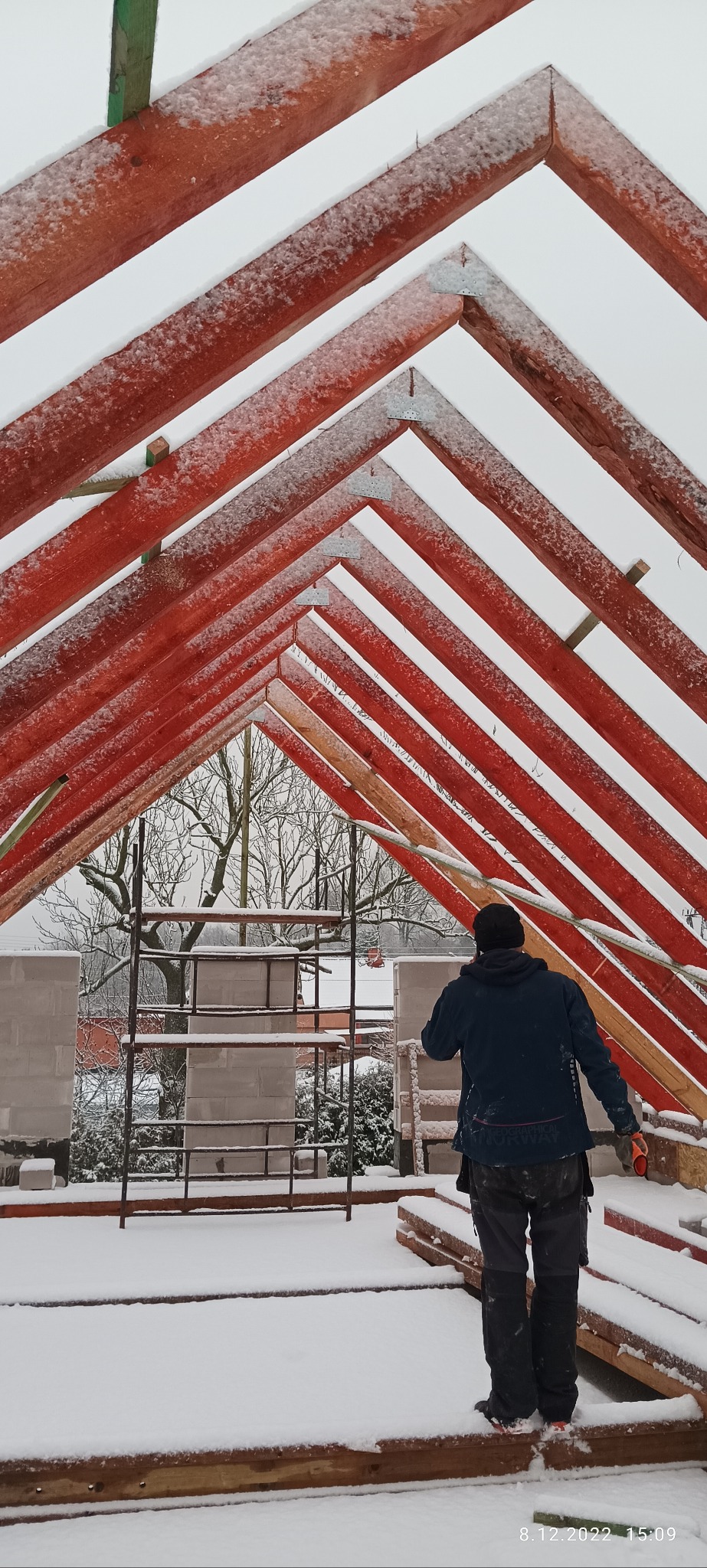 Czerwona więźba dachowa pokryta śniegiem, widok od dołu, z pracownikiem w tle i ośnieżonymi bloczkami oraz rusztowaniem w oddali.