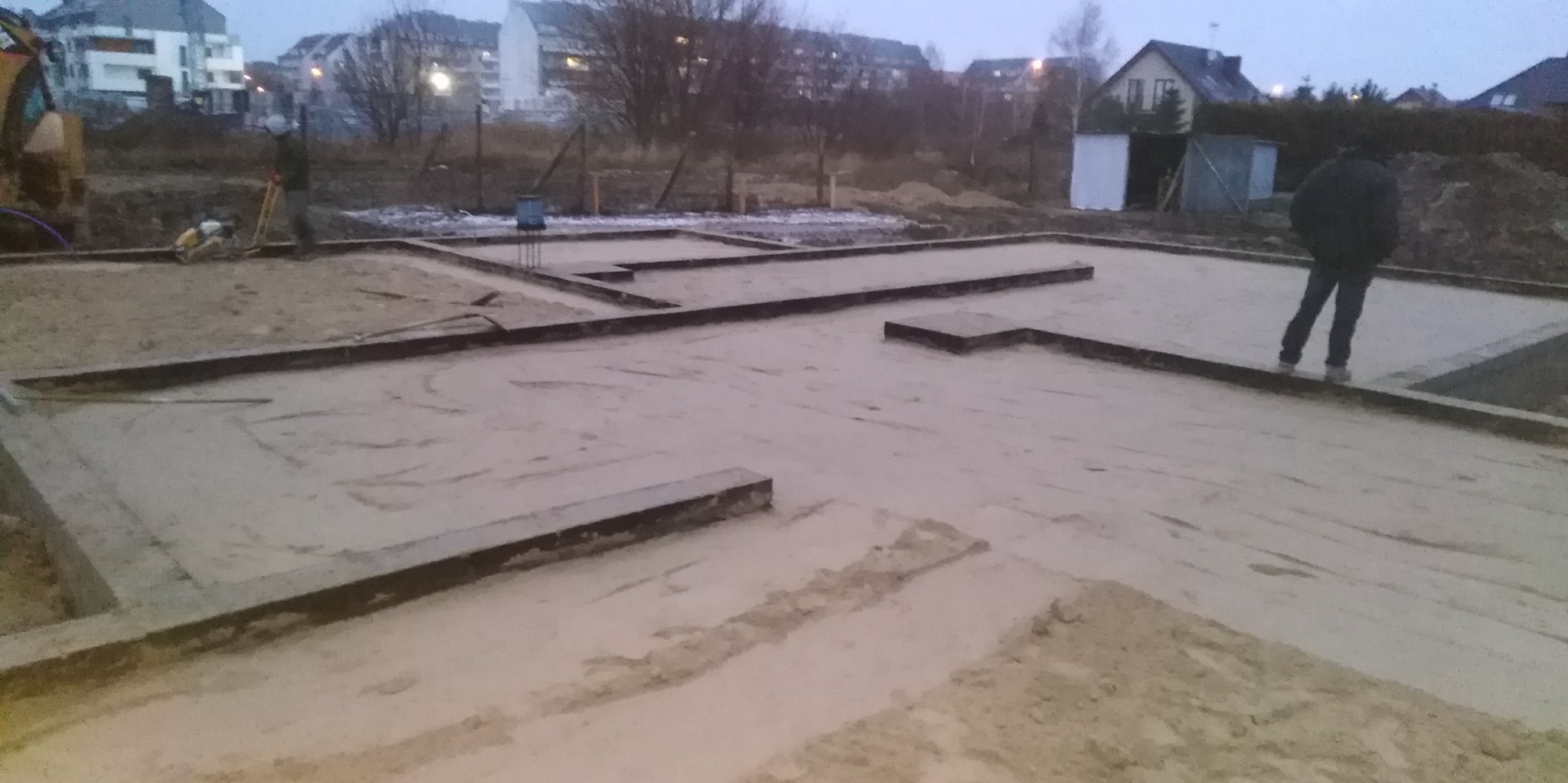 Przygotowanie gruntu pod fundamenty domu: ubijanie piasku zagęszczarką w szalunkach, w tle zabudowa jednorodzinna.