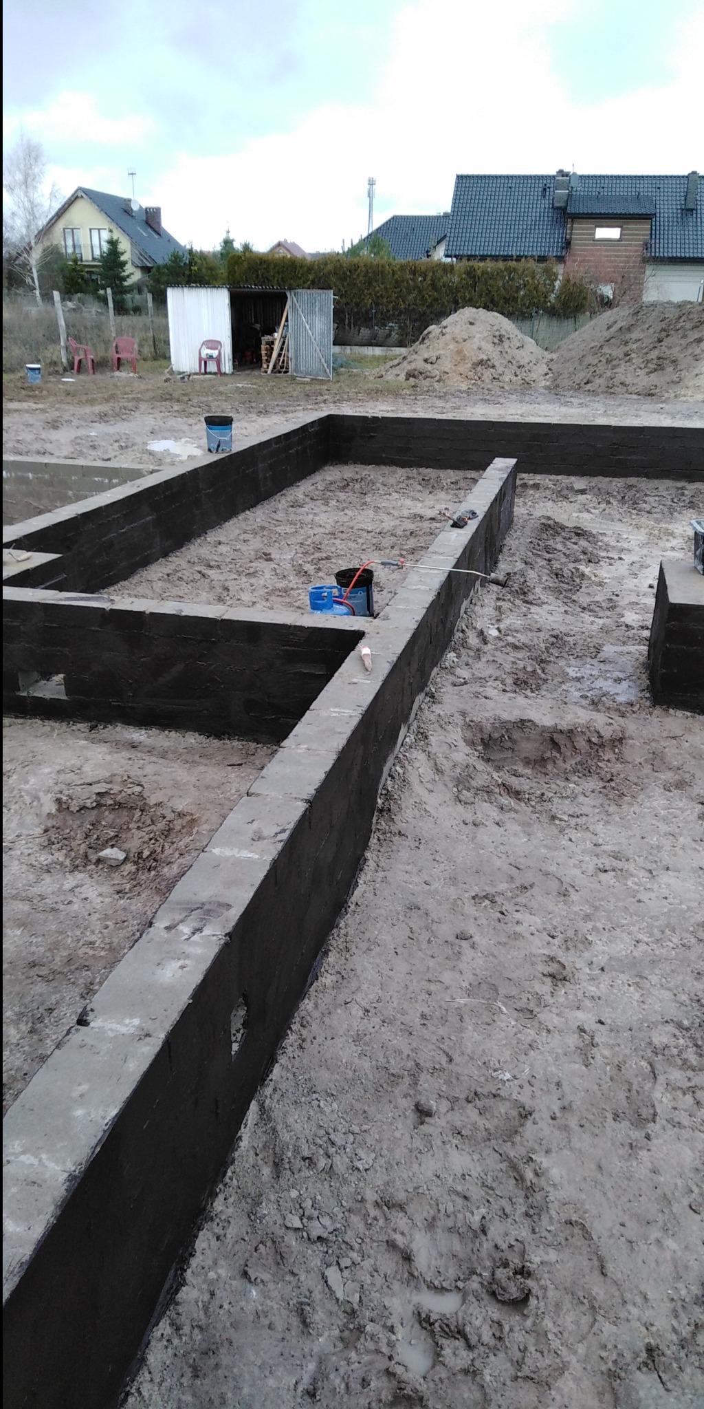 Wylane fundamenty domu z hydroizolacją pionową, widoczny teren budowy z narzędziami i pryzmą piasku w tle.