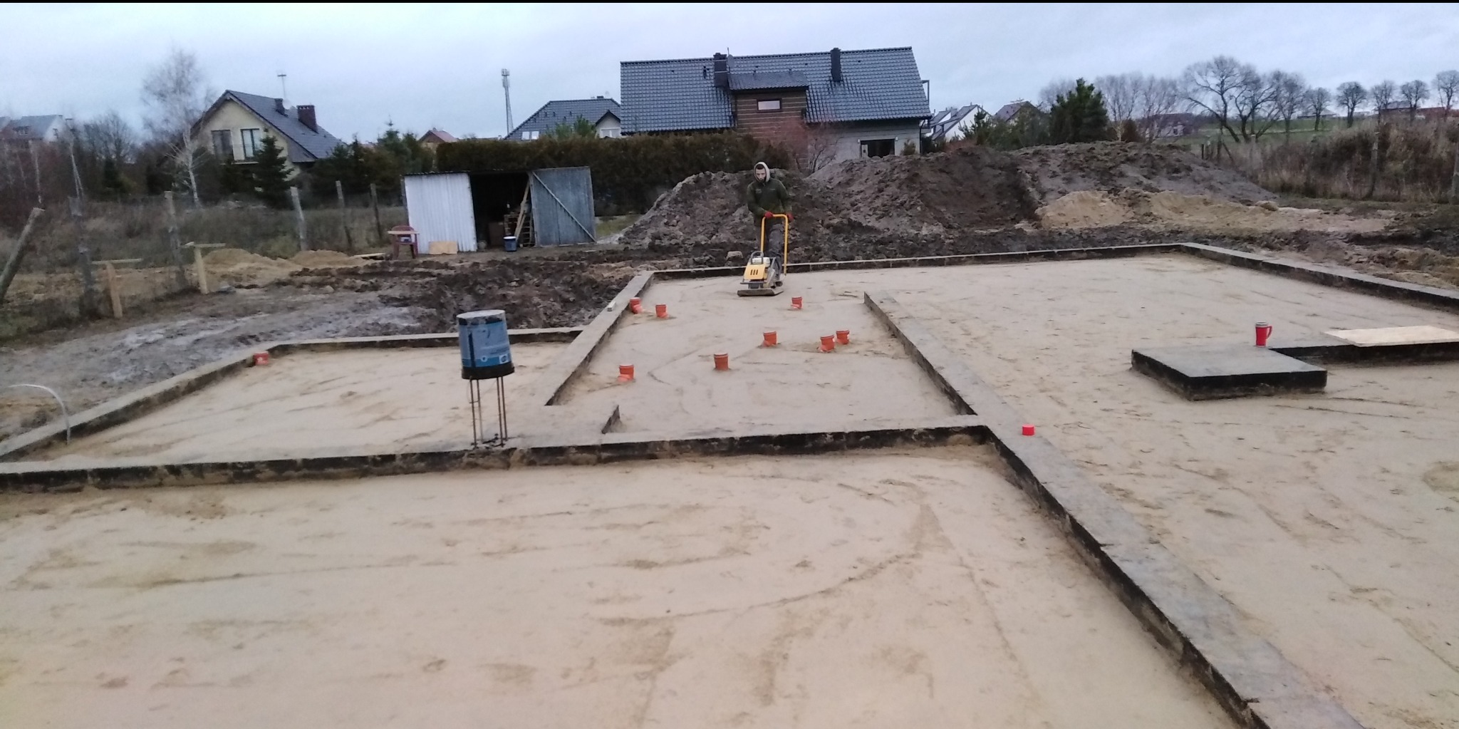 Przygotowanie terenu pod fundamenty domu: ubijanie piasku zagęszczarką płytową w wykopie z deskowaniem, widoczne czerwone znaczniki i niwelator laserowy.