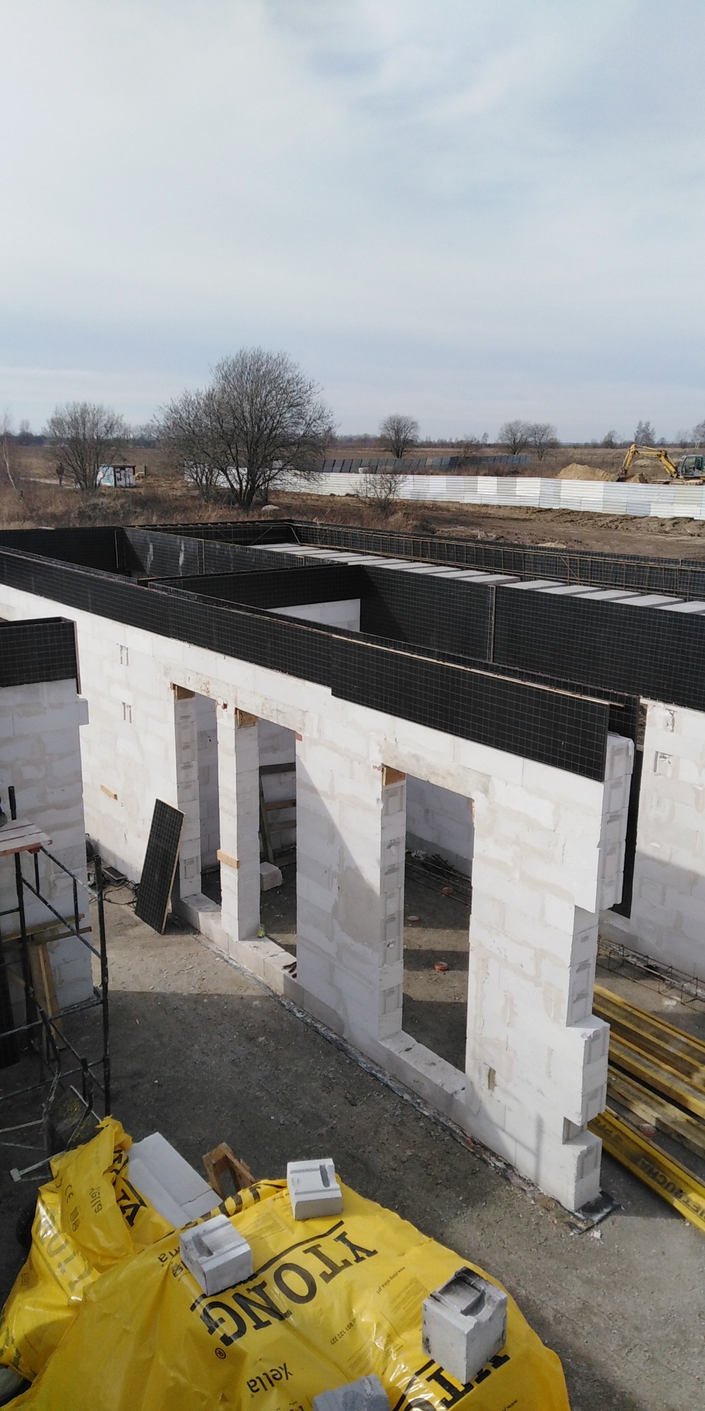 Wznoszenie ścian parteru z betonu komórkowego Ytong Silka na budowie domu, widok z góry na plac budowy z żółtą plandeką ochronną i deskami szalunkowymi w tle. W oddali krajobraz z drzewami...