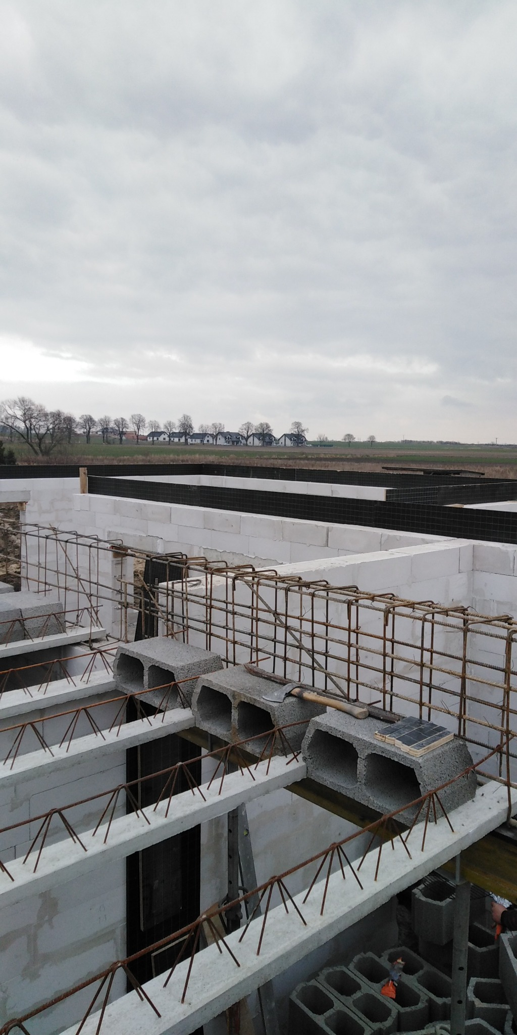 Budowa domu: Ściany fundamentowe z bloczków betonowych zbrojone stalą, widok na krajobraz z polami i zabudowaniami w tle, pochmurne niebo.