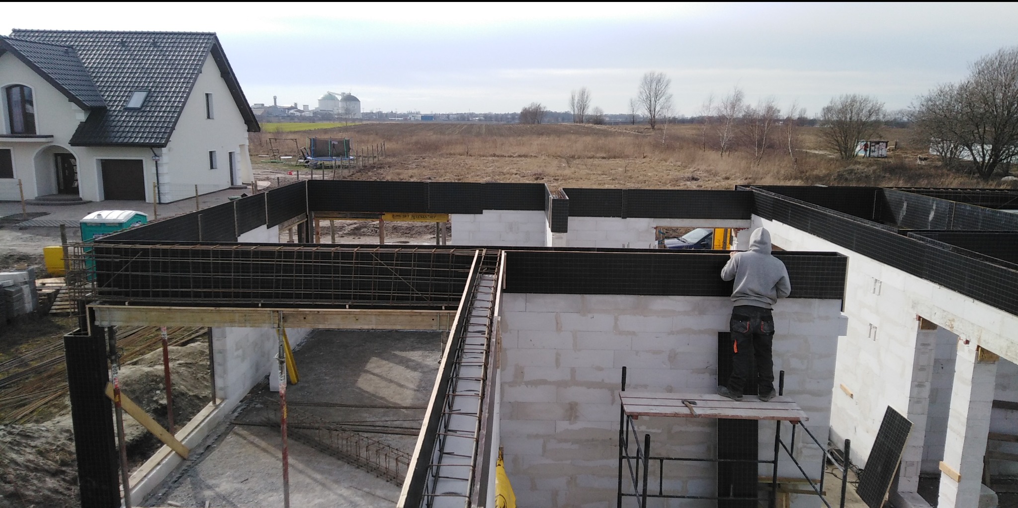 Budowa domu murowanego: pracownik na rusztowaniu montuje czarne elementy szalunku na ścianach z betonu komórkowego, w tle widoczny dom jednorodzinny i krajobraz.