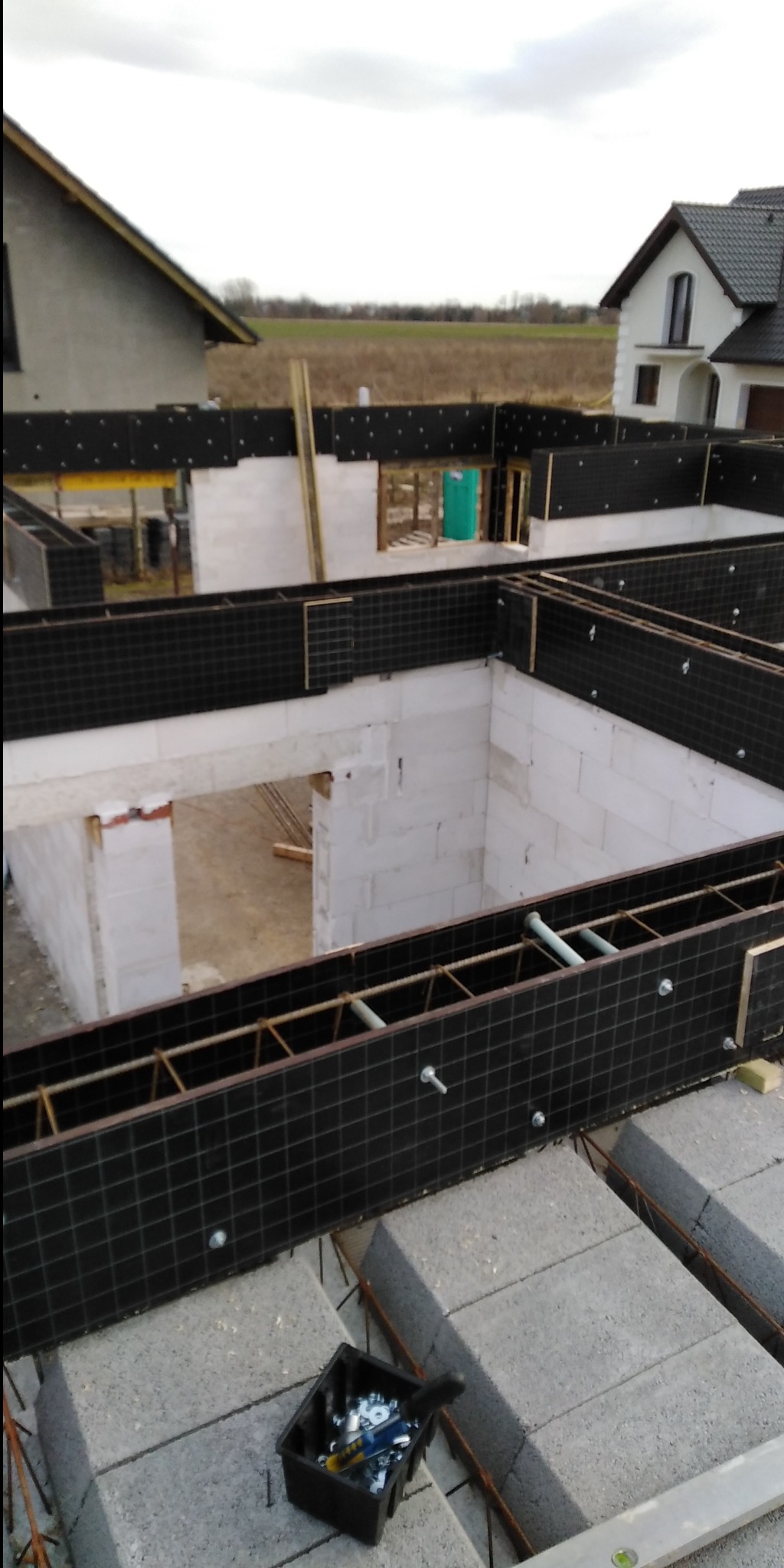 Budowa domu z bloczków betonowych: widok na fundamenty i ściany parteru z czarnym szalunkiem, narzędzia w pojemniku na pierwszym planie, w tle widoczne inne budynki i pole.