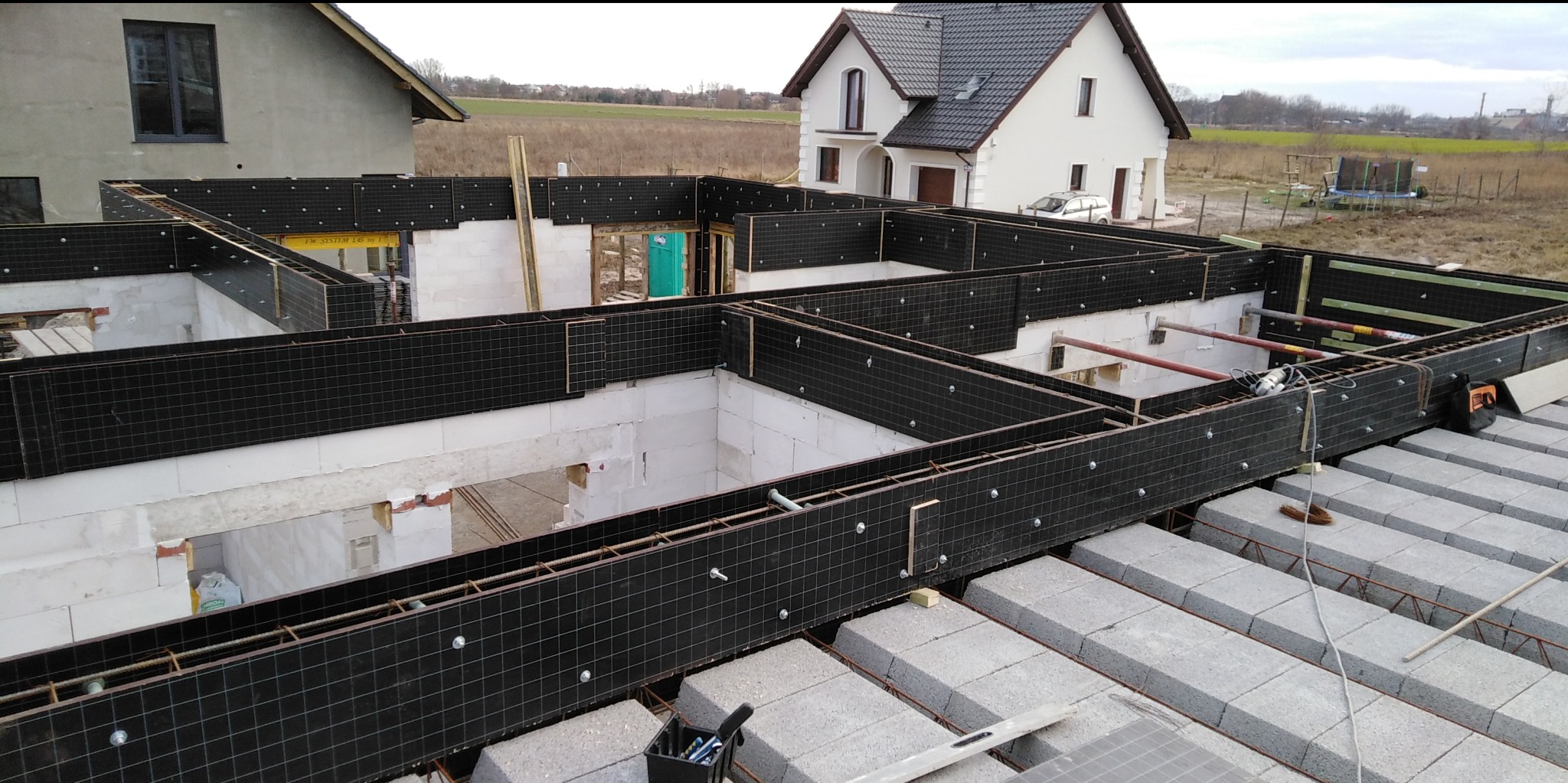 Fundamenty domu z bloczków betonowych i czarną izolacją, widok z góry na plac budowy z dwoma ukończonymi domami w tle.
