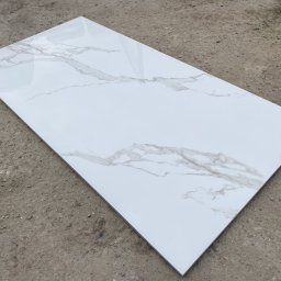 MAXI GRES - Pojedyncza, prostokątna płytka podłogowa z białego gresu polerowanego z szarym wzorem imitującym marmur, leżąca na szarym, betonowym podłożu.