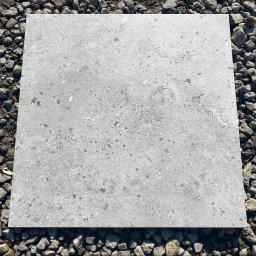 MAXI GRES - Szara, kwadratowa płyta chodnikowa z teksturą lastryko, leżąca na podłożu z drobnych kamieni.