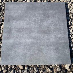 MAXI GRES - Pojedyncza, kwadratowa płyta podłogowa w odcieniach szarości z delikatną teksturą, ułożona na podłożu z drobnych kamieni.