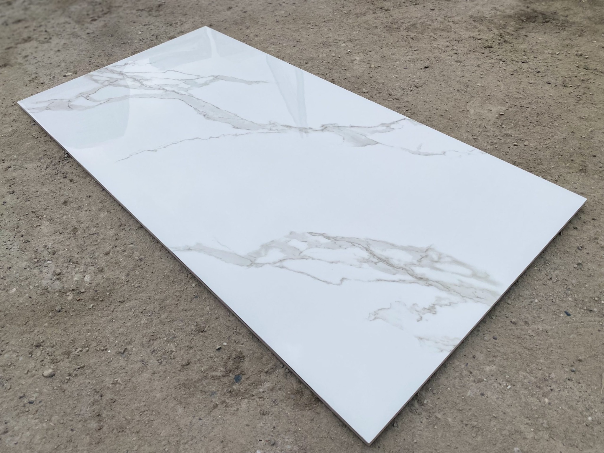 Pojedyncza, prostokątna płytka podłogowa z białego gresu polerowanego z szarym wzorem imitującym marmur, leżąca na szarym, betonowym podłożu.