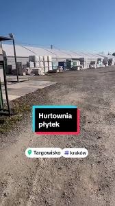 Hurtownia płytek budowlanych na zewnątrz, widoczne palety z materiałami pod zadaszeniem, w tle błękitne niebo.