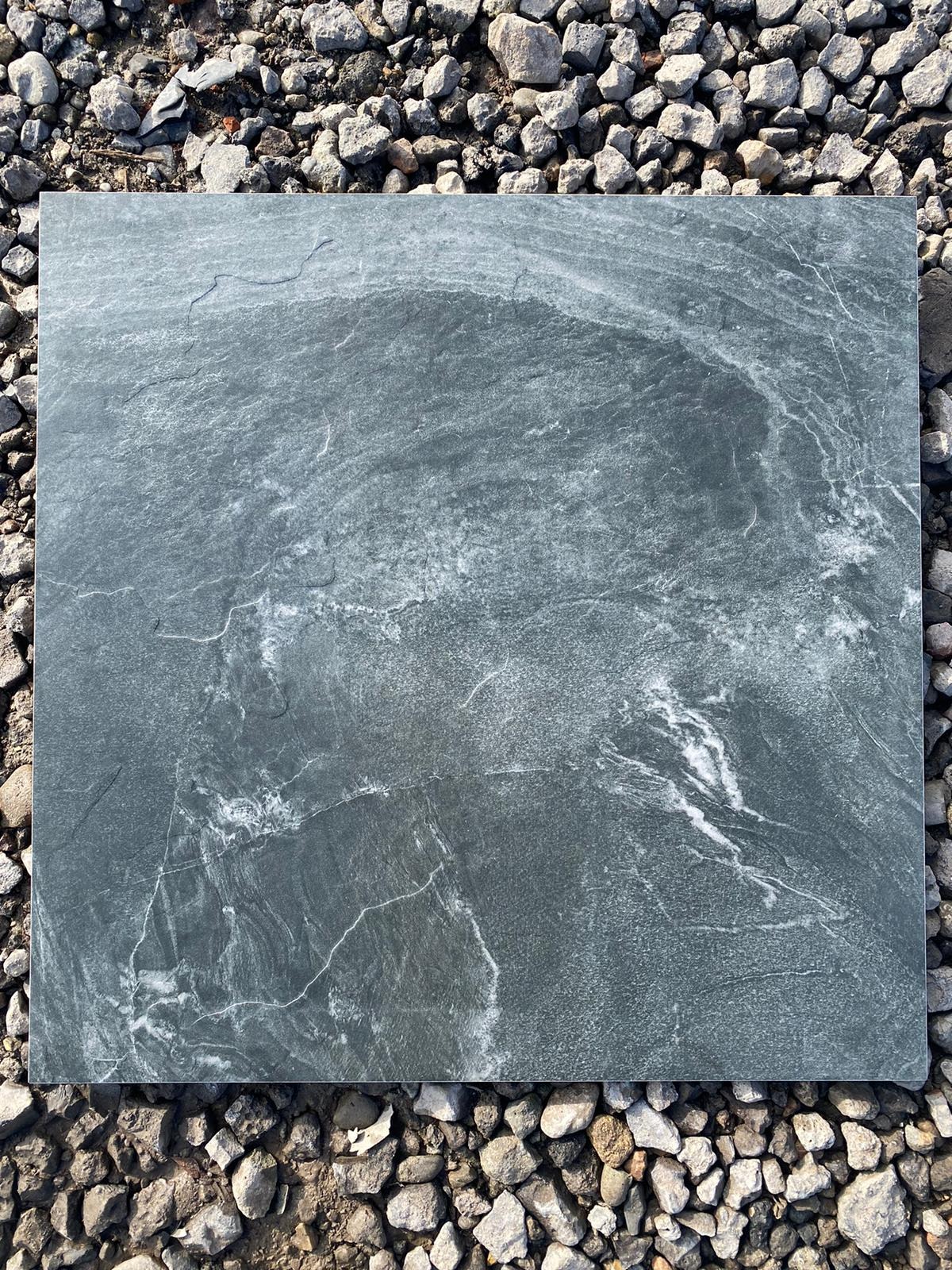 Płyta z szarego kamienia imitującego marmur, z widocznymi białymi żyłkami, leżąca na tle drobnych, szarych kamieni.
