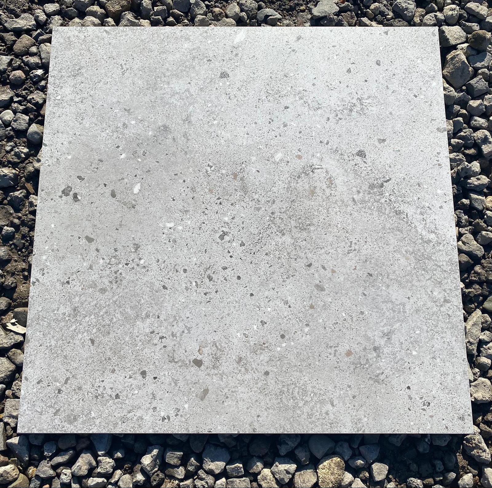 Szara, kwadratowa płyta chodnikowa z teksturą lastryko, leżąca na podłożu z drobnych kamieni.