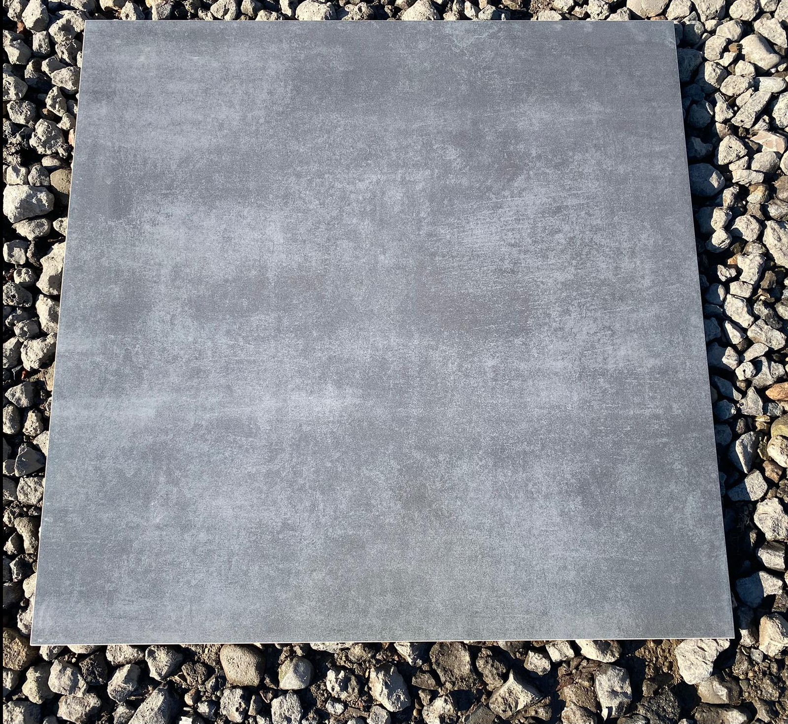 Pojedyncza, kwadratowa płyta podłogowa w odcieniach szarości z delikatną teksturą, ułożona na podłożu z drobnych kamieni.