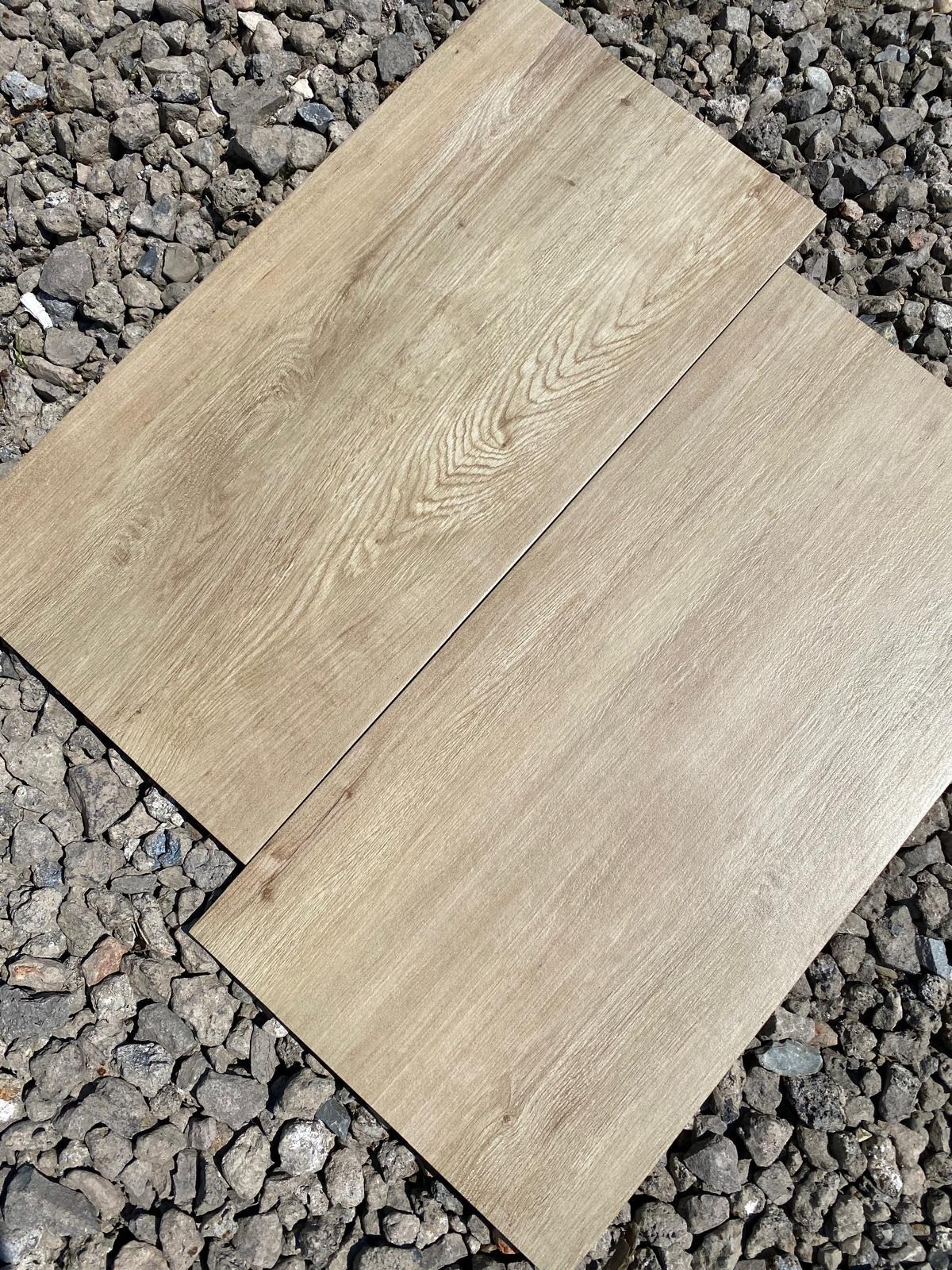 Dwa panele podłogowe o jasnym, drewnopodobnym wzorze, ułożone na tle szarego żwiru. Widoczna tekstura paneli imitująca słoje drewna.