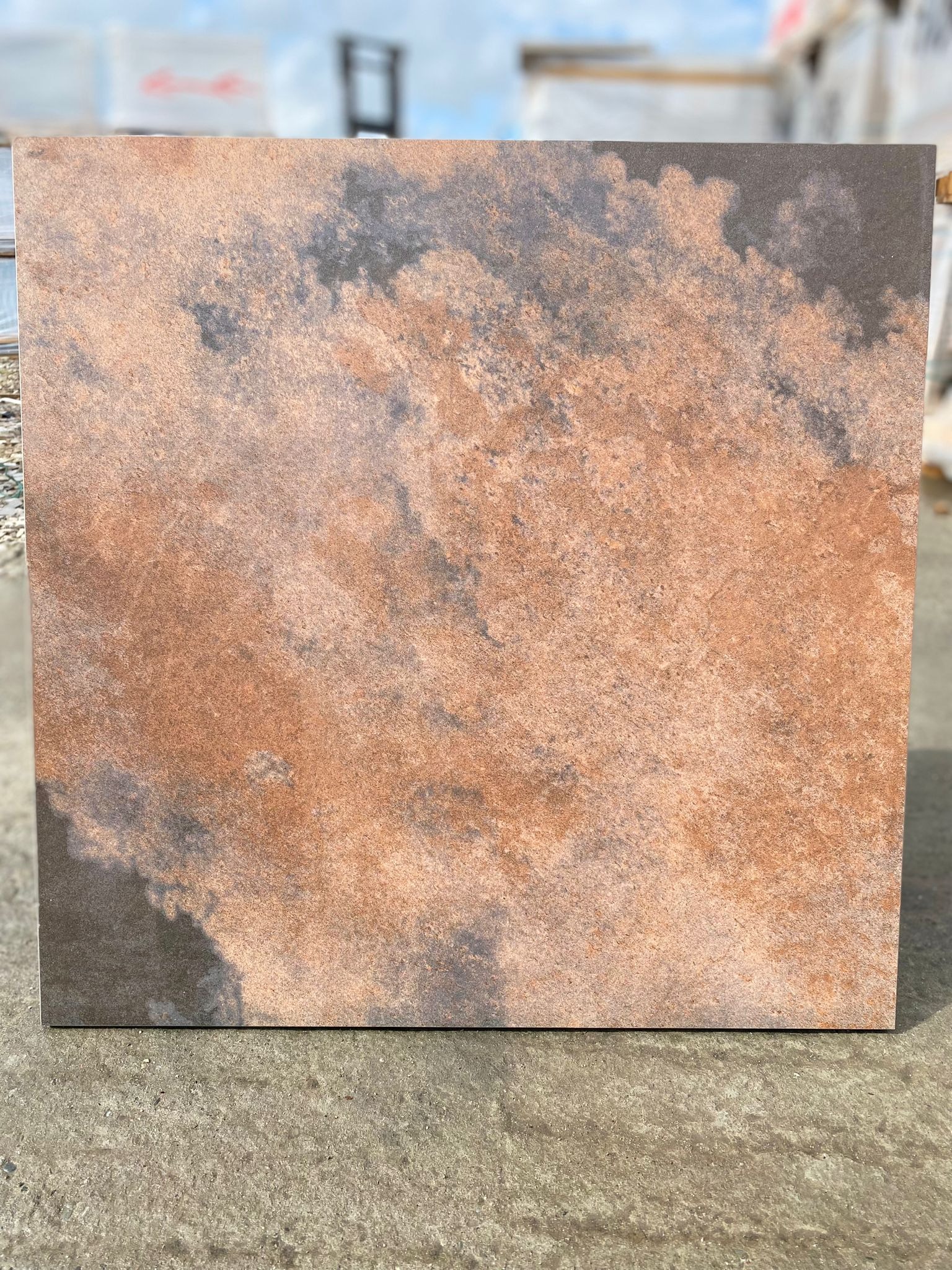 Pojedyncza płytka podłogowa o nieregularnym, rdzawym wzorze przypominającym chmurę, prezentowana na tle betonowej powierzchni w świetle dziennym.