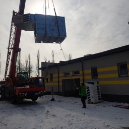 Marek Felskowski Coldest Group - Montaż centrali wentylacyjnej VBW za pomocą dźwigu na zewnątrz budynku w zimowy dzień, pracownik asekuruje liną.