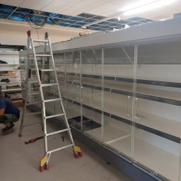 Marek Felskowski Coldest Group - Montaż chłodniczej lady sklepowej: pracownik w kucki, aluminiowa drabina z żółtymi nakładkami, pusta lada z szklanymi drzwiami i półkami, widoczne narzędzia, sufit z widocznymi kablami.