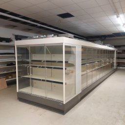 Marek Felskowski Coldest Group - Długa, pusta chłodnicza lada sklepowa z przeszklonymi drzwiami i półkami, ustawiona w surowym wnętrzu sklepu, z widocznymi elementami instalacji pod sufitem.