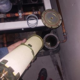 Marek Felskowski Coldest Group - Filtr cylindryczny z metalowymi elementami w dłoni, obok rozmontowanego fragmentu instalacji z rurami miedzianymi i betonową podłogą.