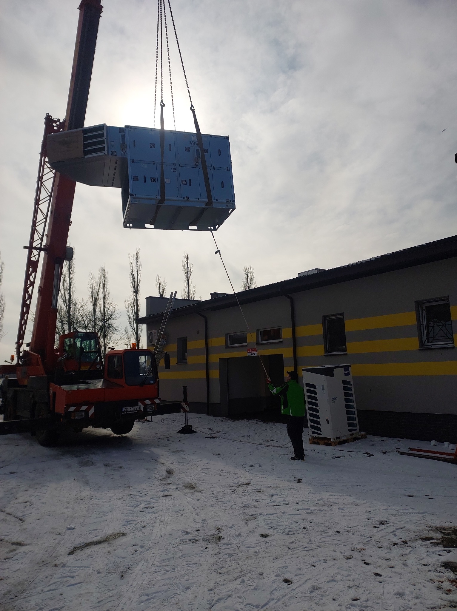 Montaż centrali wentylacyjnej VBW za pomocą dźwigu na zewnątrz budynku w zimowy dzień, pracownik asekuruje liną.