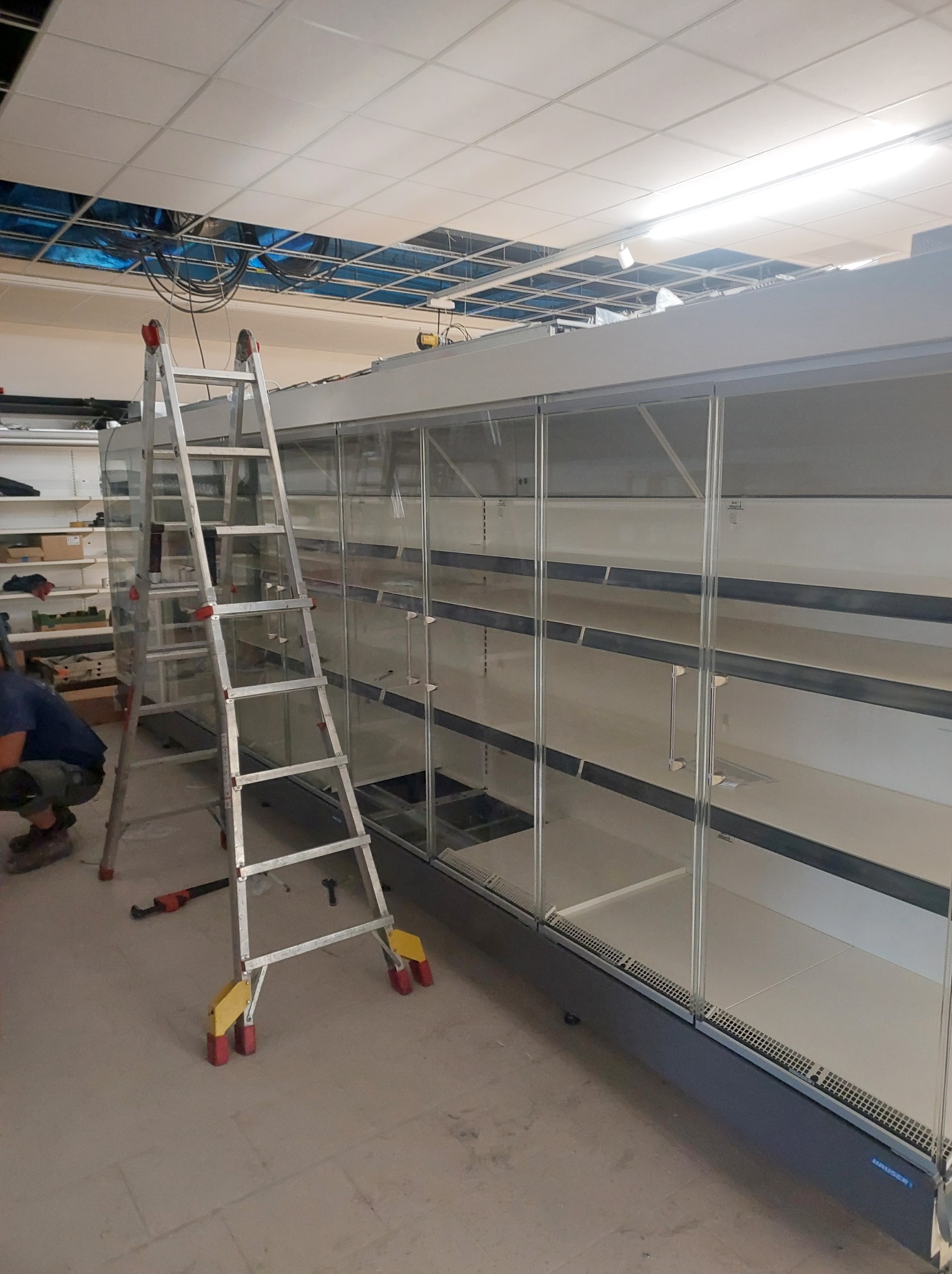 Montaż chłodniczej lady sklepowej: pracownik w kucki, aluminiowa drabina z żółtymi nakładkami, pusta lada z szklanymi drzwiami i półkami, widoczne narzędzia, sufit z widocznymi kablami.