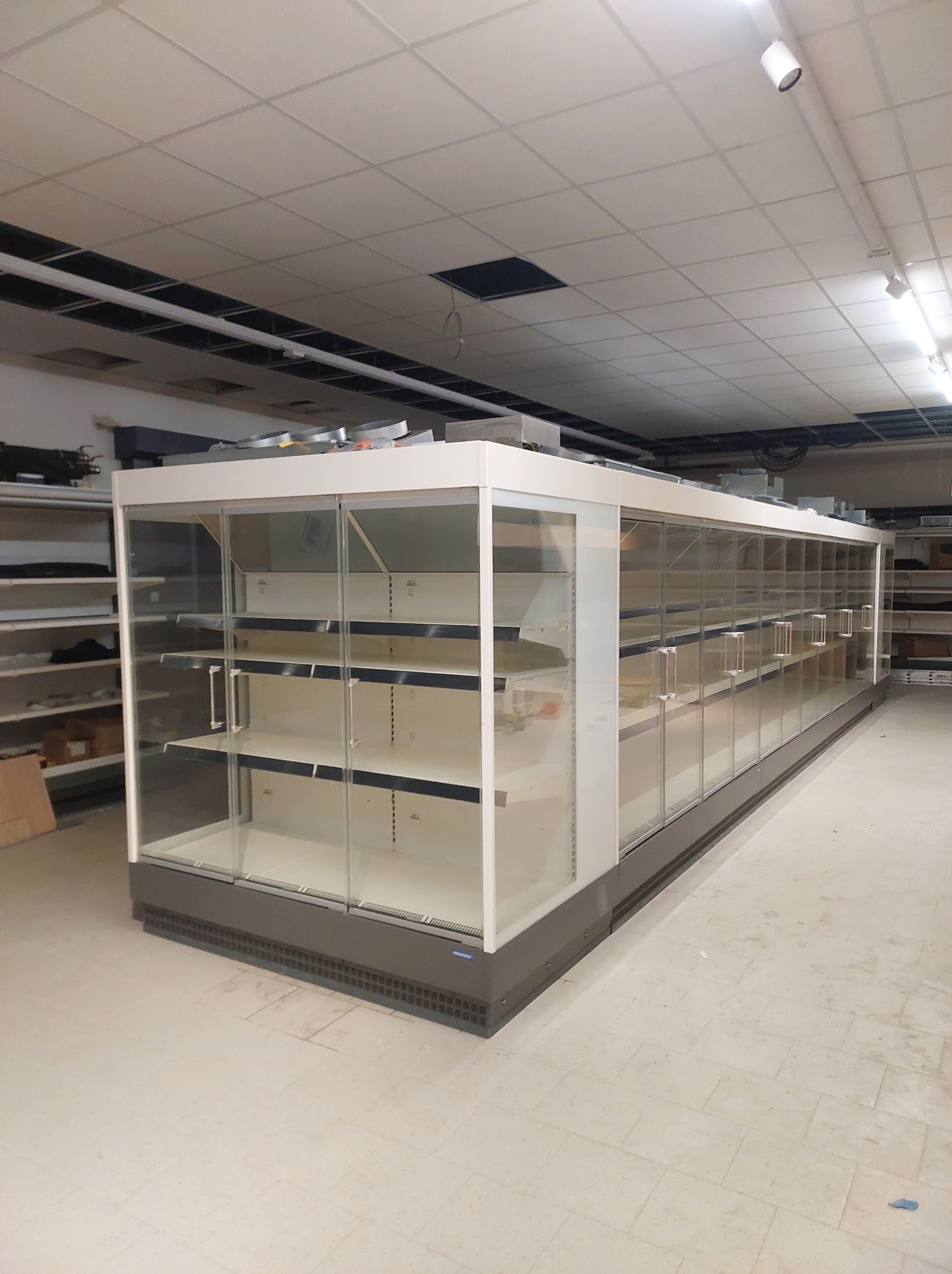 Długa, pusta chłodnicza lada sklepowa z przeszklonymi drzwiami i półkami, ustawiona w surowym wnętrzu sklepu, z widocznymi elementami instalacji pod sufitem.