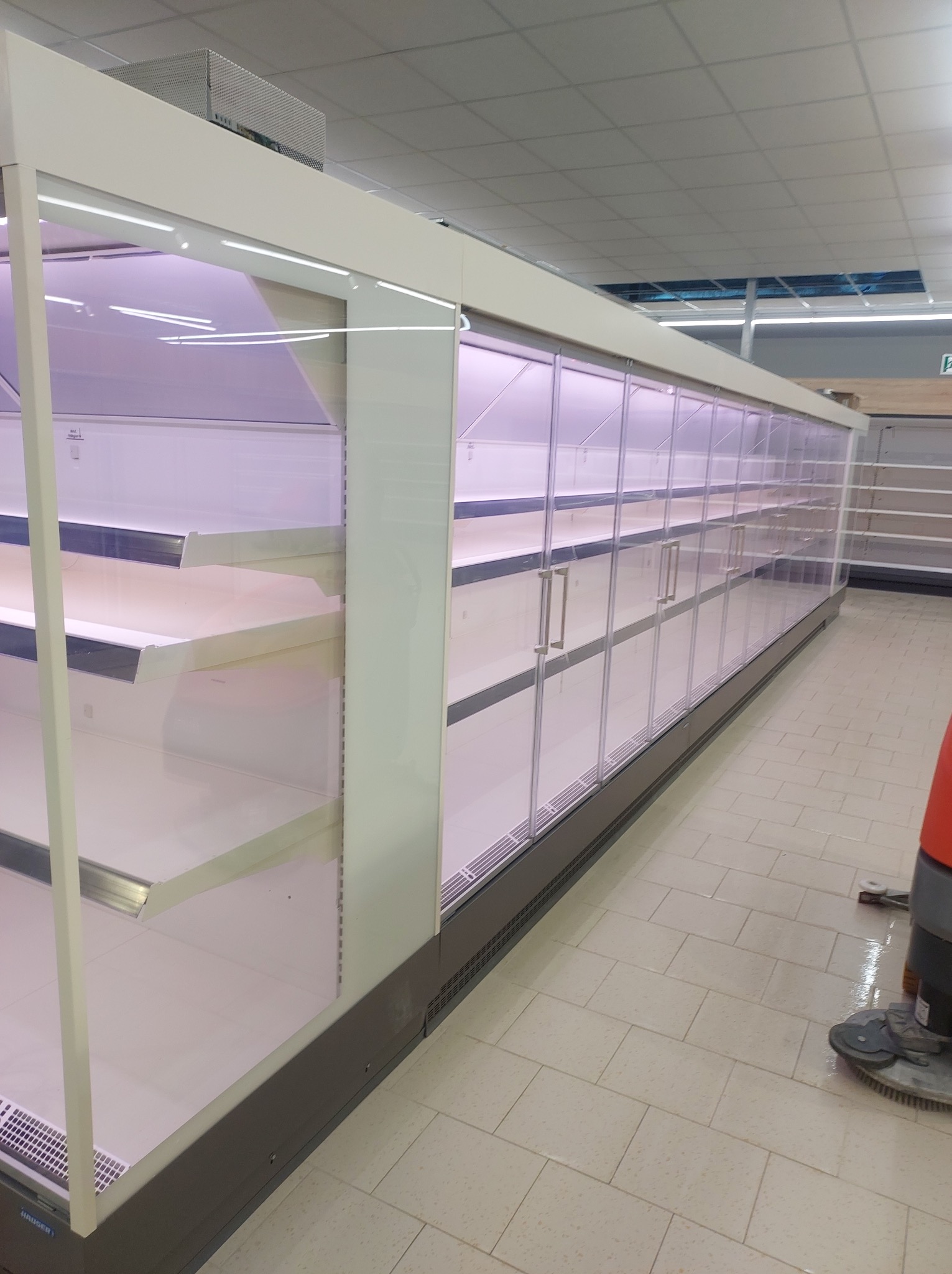 Długa, pusta chłodnicza witryna sklepowa z przesuwnymi szklanymi drzwiami i fioletowym oświetleniem, częściowo widoczna maszyna czyszcząca podłogę.