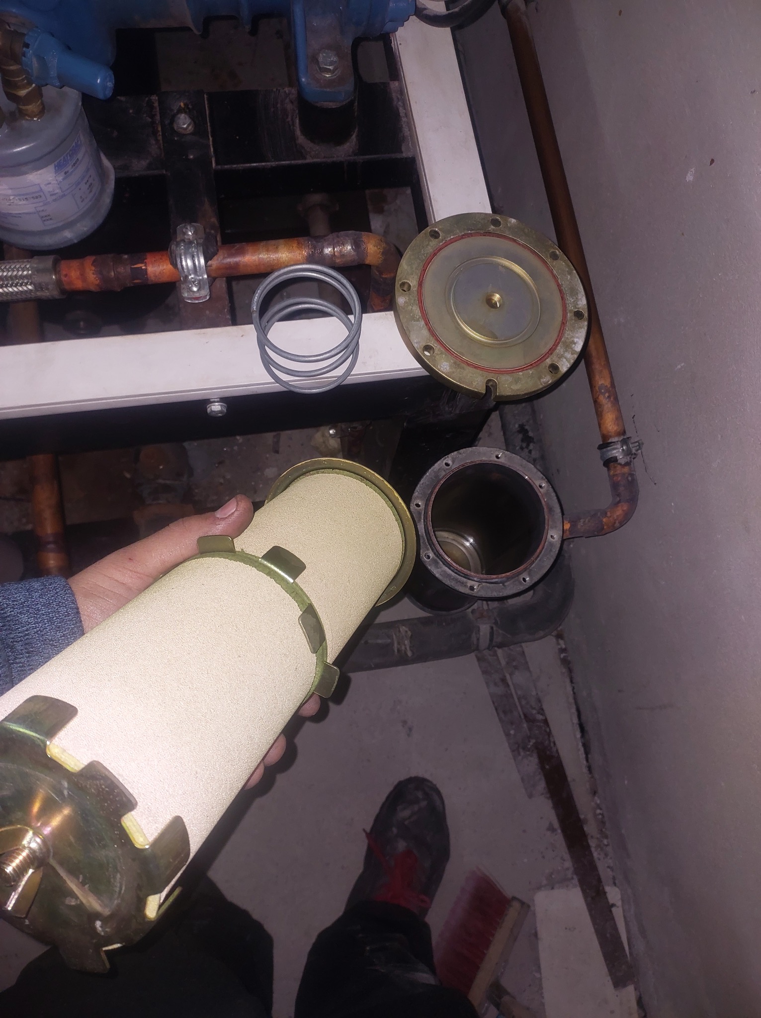 Filtr cylindryczny z metalowymi elementami w dłoni, obok rozmontowanego fragmentu instalacji z rurami miedzianymi i betonową podłogą.