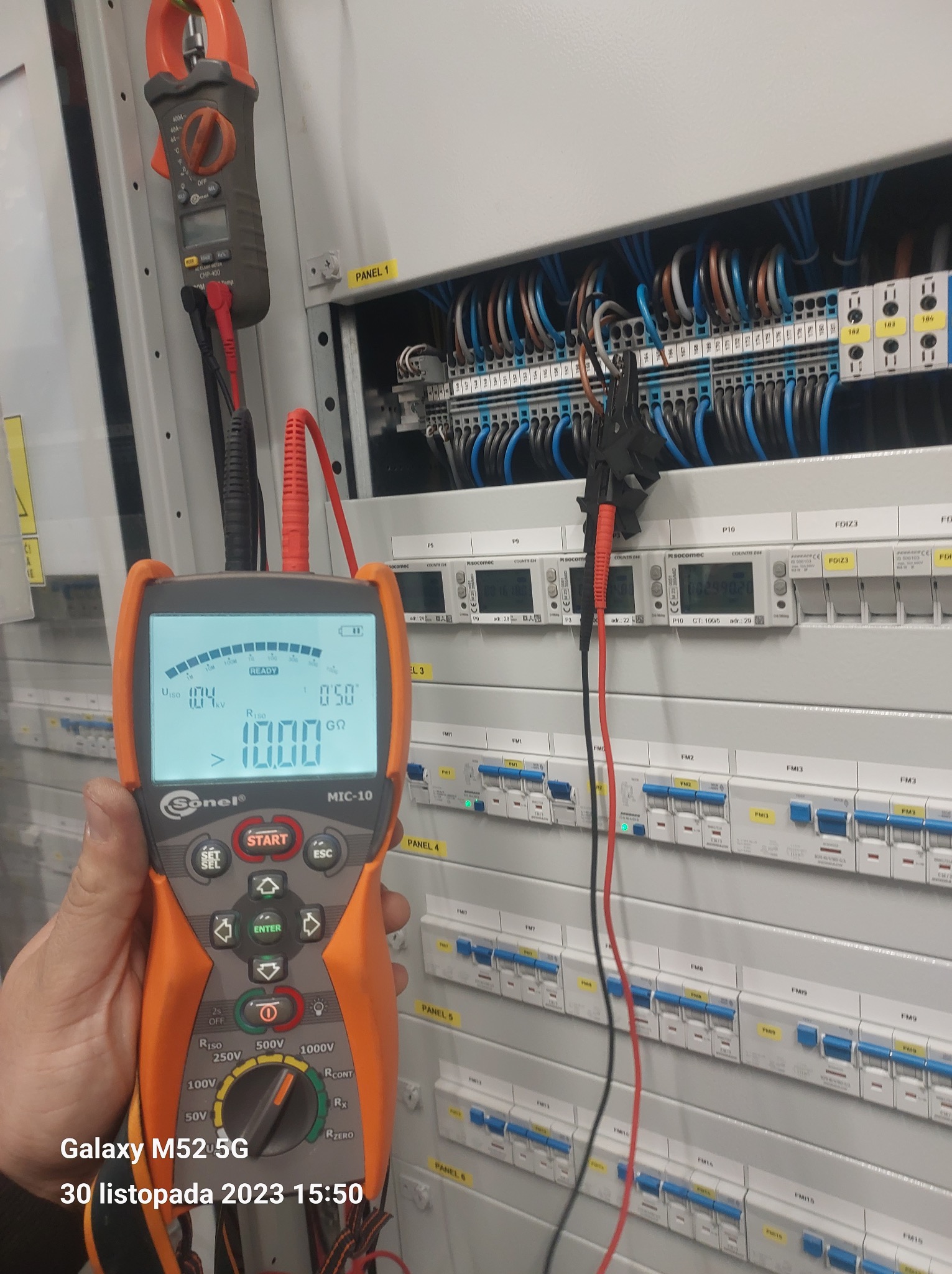 Elektryk mierzy rezystancję izolacji w rozdzielnicy elektrycznej za pomocą miernika Sonel MIC-10, widoczne bezpieczniki i okablowanie.
