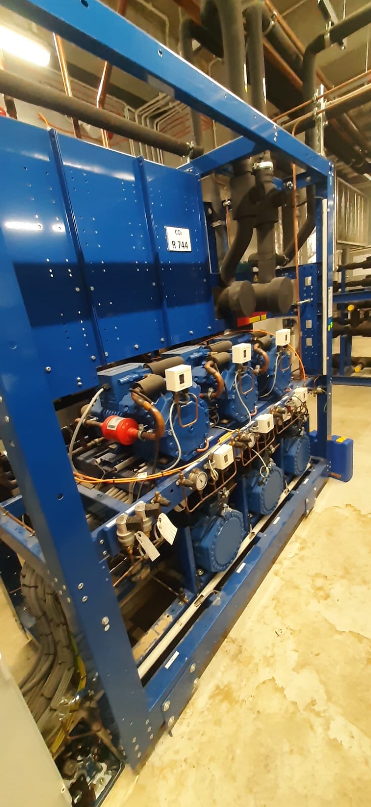 Niebieska rama z czterema sprężarkami chłodniczymi, widoczne rury miedziane, zawory i manometry, etykieta z napisem CO2 R.744, instalacja w pomieszczeniu technicznym.