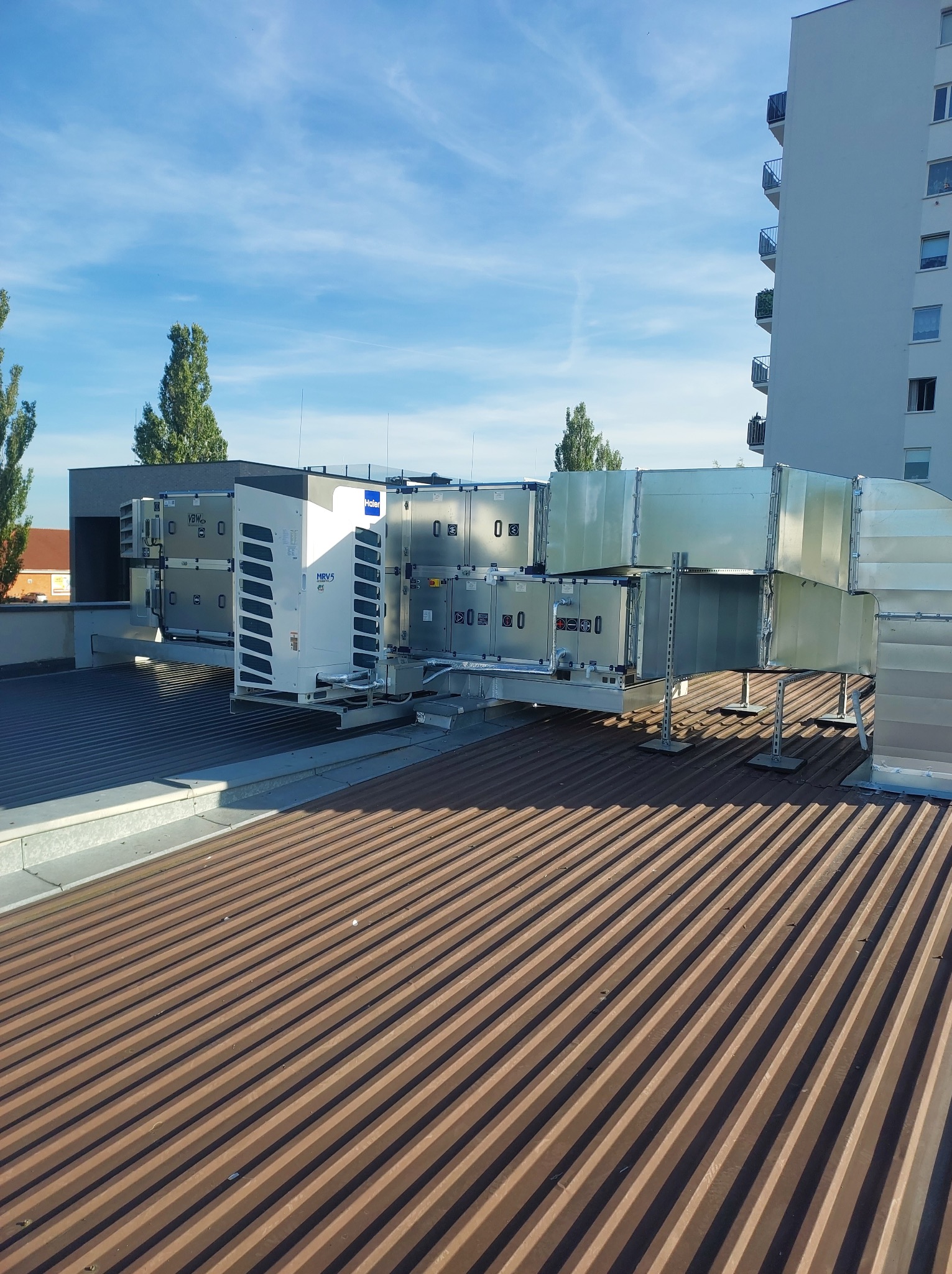 Instalacja wentylacyjna na dachu budynku mieszkalnego, widoczne jednostki zewnętrzne i kanały wentylacyjne, błękitne niebo w tle.