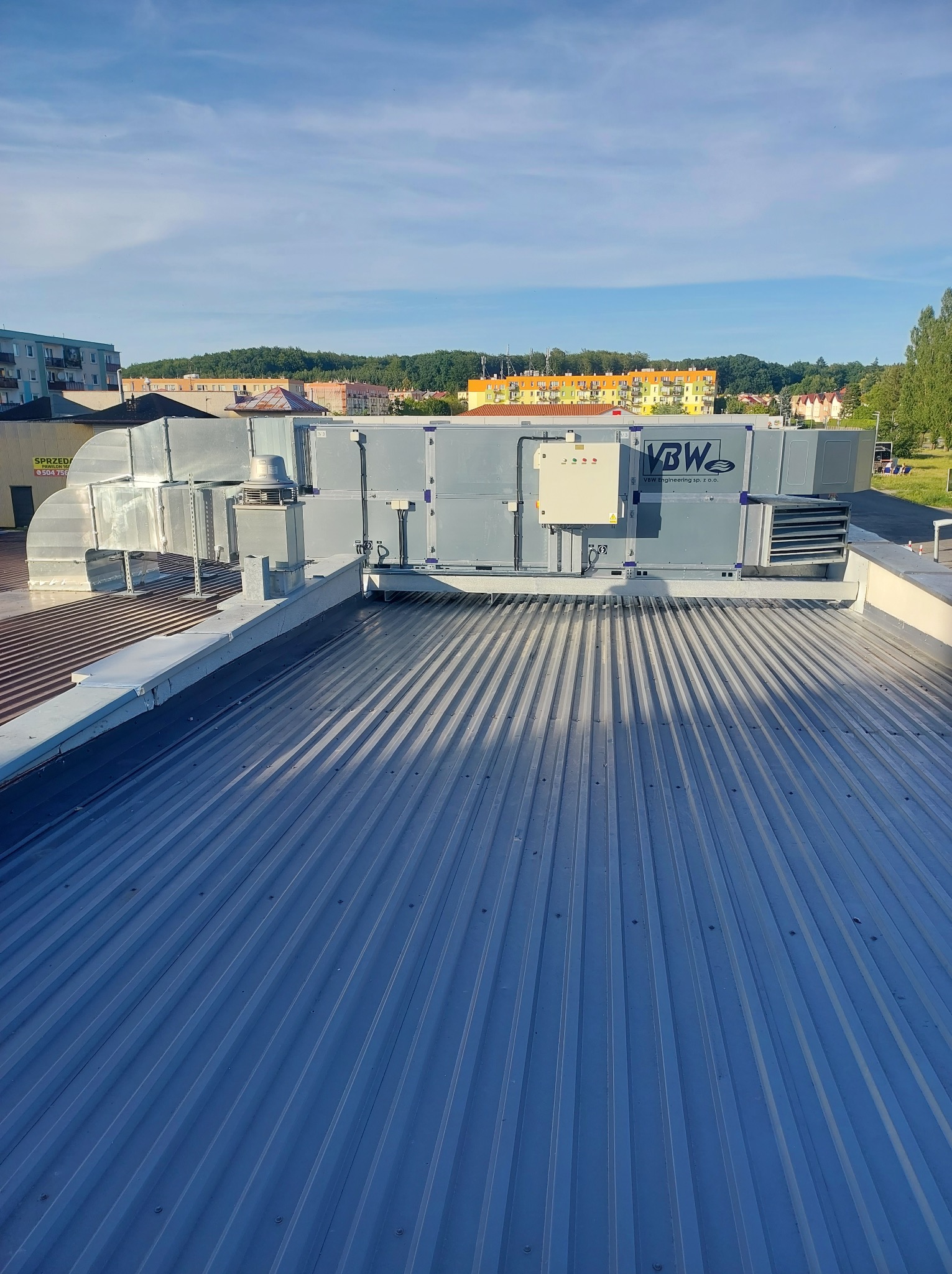 Widok z góry na instalację wentylacyjną z centralą na dachu budynku, z widocznymi przewodami wentylacyjnymi i elementami sterującymi, na tle zabudowy miejskiej i błękitnego nieba.
